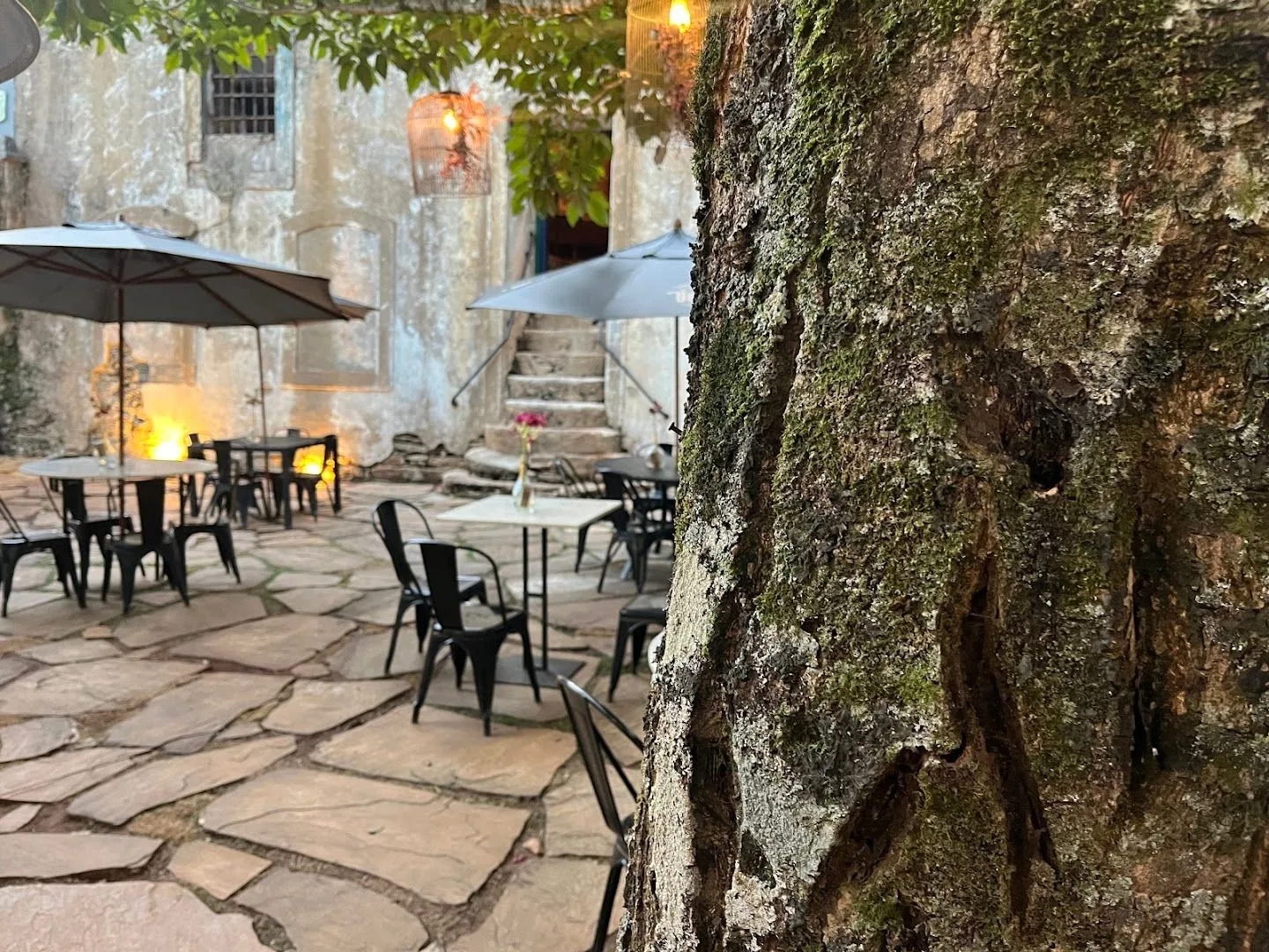 Pátio de pedra com mesas, guarda-sóis e lampiões acesos ao entardecer